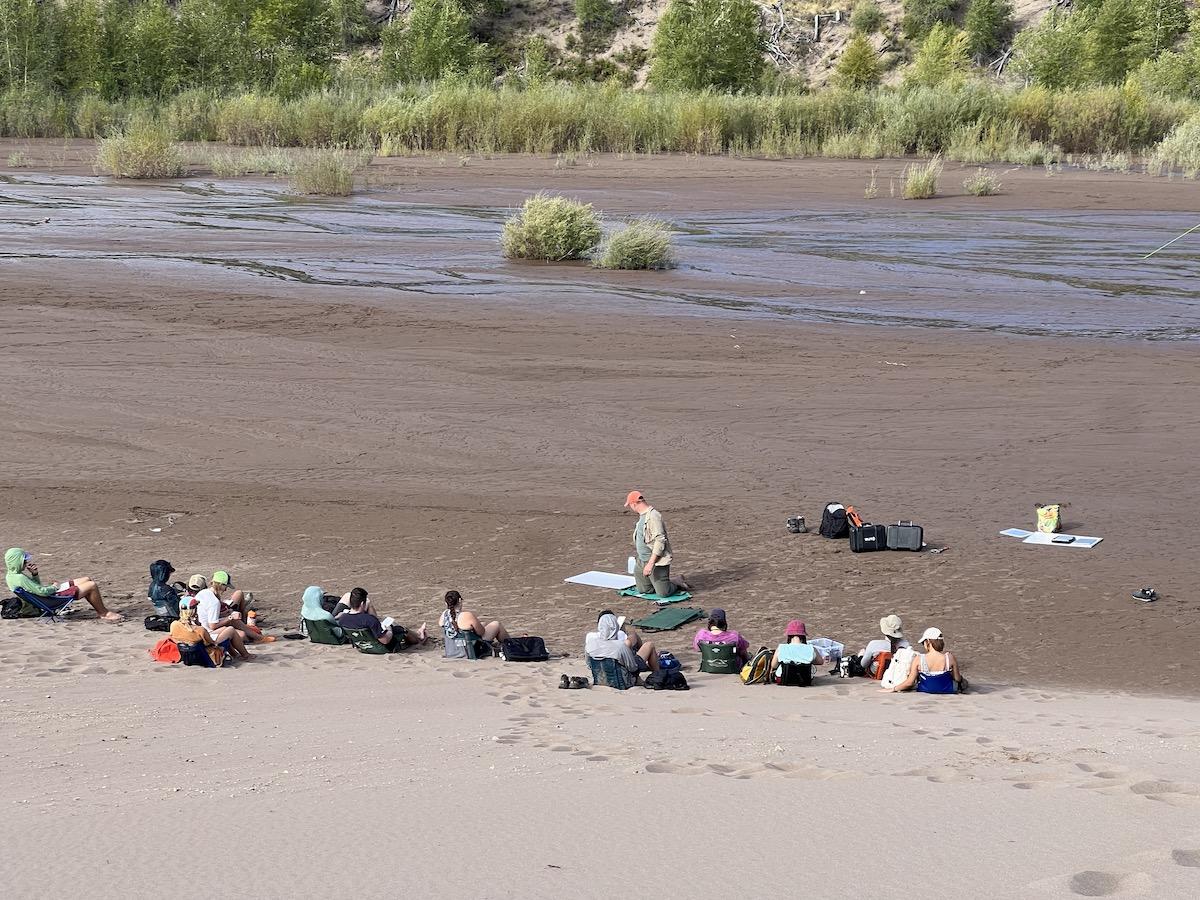 一群学生坐在沙滩上，身后是一条小溪，他们正在研究河岸地带